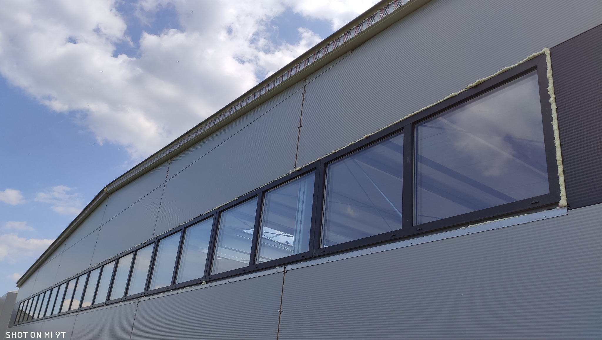 Rząd czarnych okien w metalowej hali przemysłowej, uszczelnionych pianką montażową, widok z dołu na błękitne niebo z chmurami.