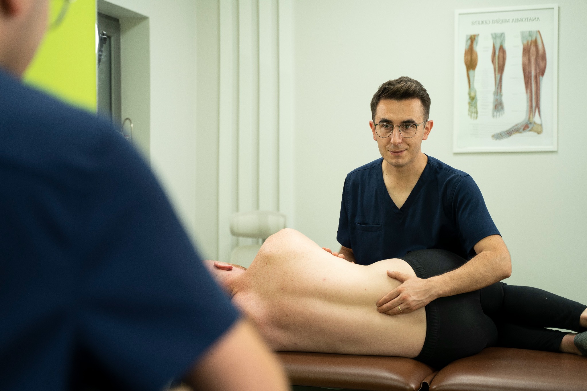 Mężczyzna w okularach, w granatowym uniformie, wykonuje badanie palpacyjne pleców pacjenta leżącego na brązowym stole do masażu w gabinecie z plakatem anatomicznym.