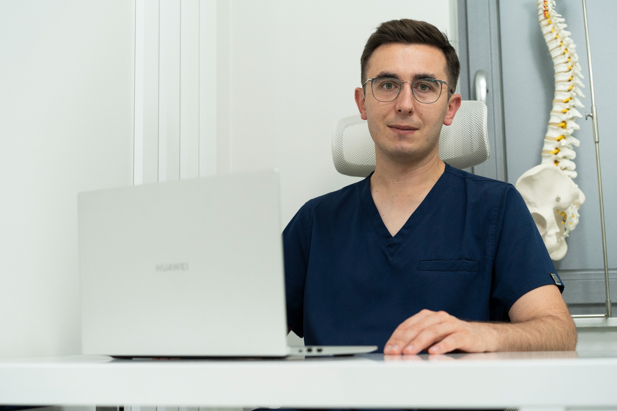 Lekarz w okularach siedzi za biurkiem z laptopem Huawei, w tle model kręgosłupa.