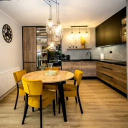 Nowoczesna kuchnia z drewnianymi frontami i czarnymi elementami, okrągły stół z żółtymi krzesłami, designerskie lampy wiszące i dekoracyjny zegar na ścianie.