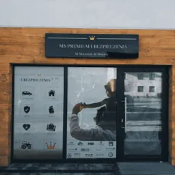 Fasada biura MS Premium Ubezpieczenia z grafikami w oknach przedstawiającymi ikony: samochodu, domu, tarczy, grupy ludzi, serca, walizki, traktora i korony. Na szybie odbija się sylwetka mężczyzny...