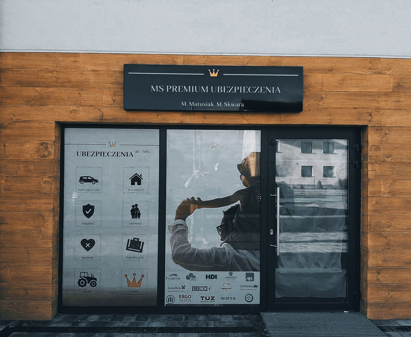 Fasada biura MS Premium Ubezpieczenia z grafikami w oknach przedstawiającymi ikony: samochodu, domu, tarczy, grupy ludzi, serca, walizki, traktora i korony. Na szybie odbija się sylwetka mężczyzny...