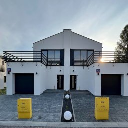 Build Apro sp.z o o - Nowoczesny bliźniak z białymi elewacjami, czarnymi balustradami balkonów i bramami garażowymi, oświetlony popołudniowym słońcem, z żółtymi skrzynkami gazowymi na pierwszym planie.