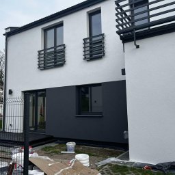 Build Apro sp.z o o - Dwukondygnacyjny dom z biało-grafitową elewacją i czarnymi ramami okiennymi, widoczne wiaderka z farbą i zabezpieczenia na trawie, sugerujące świeżo zakończone prace malarskie.