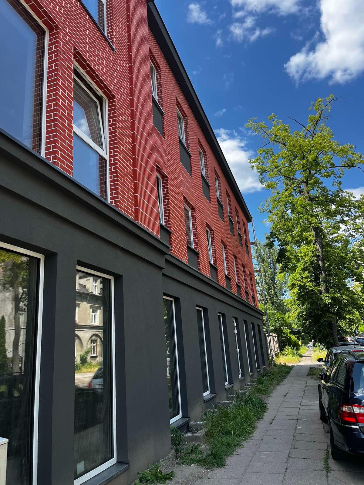 Odnowiona elewacja budynku z czerwonej cegły z szarą, gładką dolną częścią, widoczne okna z białymi ramami, chodnik i zieleń wzdłuż budynku, błękitne niebo z chmurami.