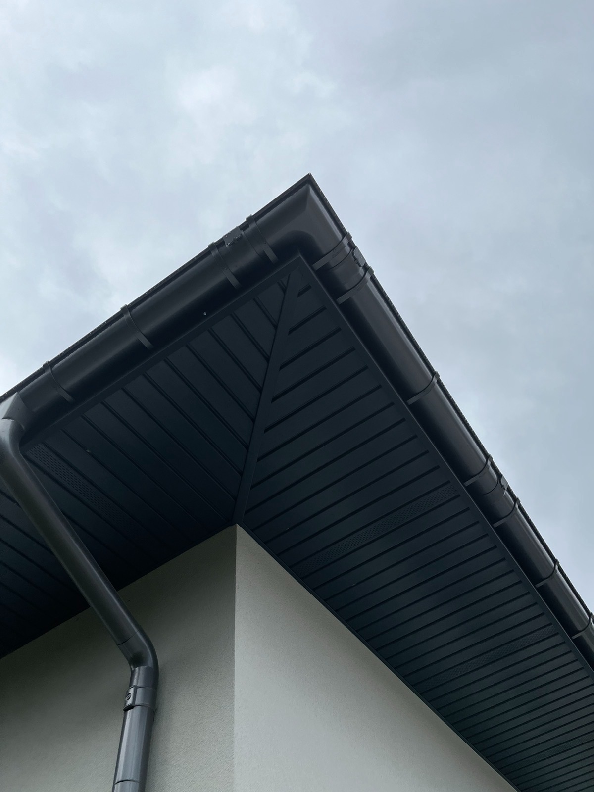 Fragment elewacji budynku z ciemną podbitką dachową, rynną i jasnym tynkiem, sfotografowany pod kątem w pochmurny dzień.