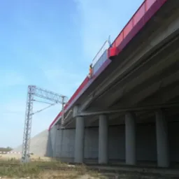 Pracownik w pomarańczowym kombinezonie na krawędzi nowo pomalowanego na czerwono wiaduktu kolejowego, widok z dołu na betonowe filary i trakcję elektryczną.