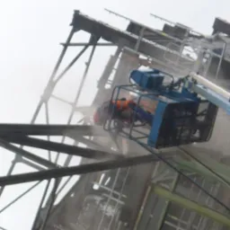 Pracownik w podnośniku koszowym czyści metalową konstrukcję z rdzy, widoczne efekty piaskowania. Ujęcie z dołu, mgła w tle.