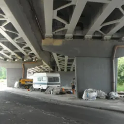 Prace konserwacyjne pod betonowym wiaduktem. Widoczny sprzęt, agregat, przyczepa kempingowa i robotnik w pomarańczowych spodniach. Częściowo odnowiona powierzchnia.