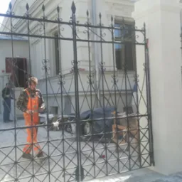 Pracownik w pomarańczowych spodniach montuje ozdobną, czarną bramę w ogrodzeniu posesji z jasną elewacją, widoczne narzędzia i sprzęt montażowy.