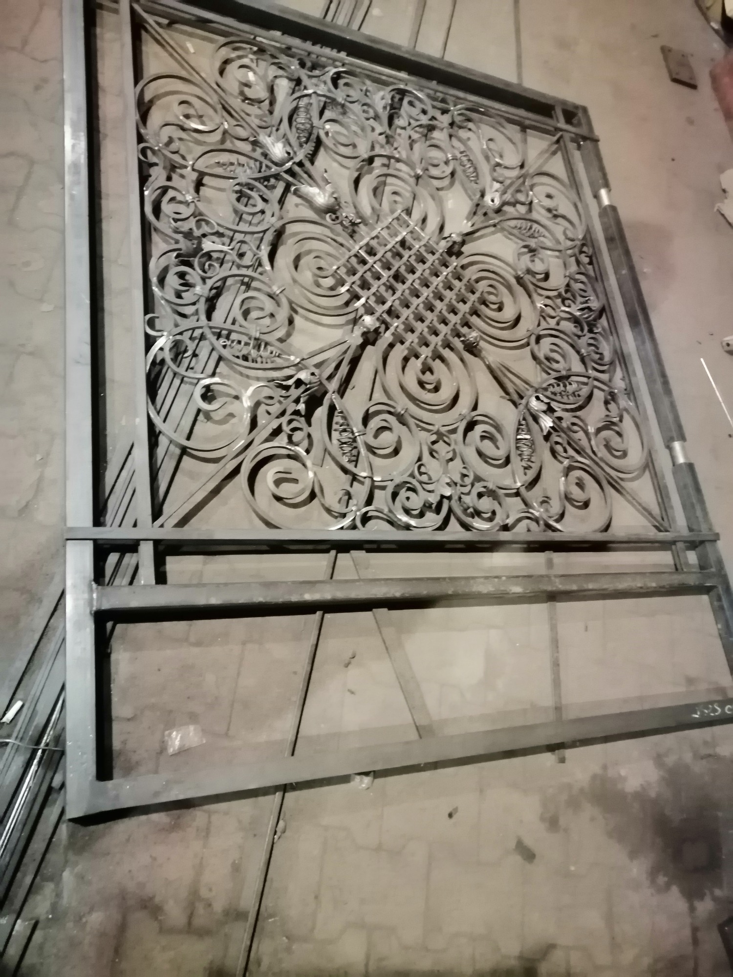 Surowy, metalowy fragment ogrodzenia z ażurowym, zdobionym wypełnieniem w warsztacie, widoczne ślady obróbki.