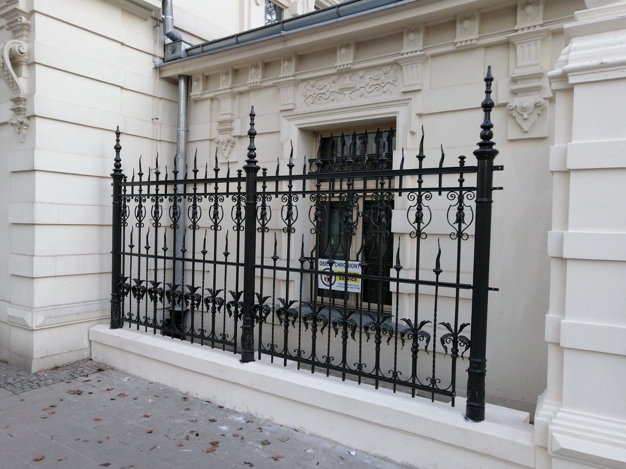 Czarny, metalowy, ozdobny płot z kutego żelaza, zamontowany na niskim, białym murku, przed budynkiem z kremową elewacją i dekoracyjnymi detalami architektonicznymi.