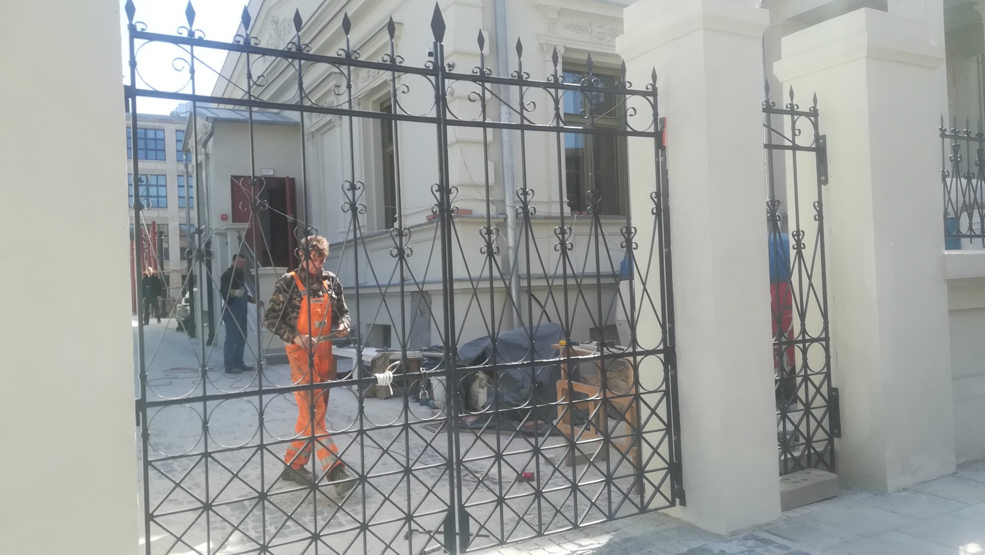 Pracownik w pomarańczowych spodniach montuje ozdobną, czarną bramę w ogrodzeniu posesji z jasną elewacją, widoczne narzędzia i sprzęt montażowy.