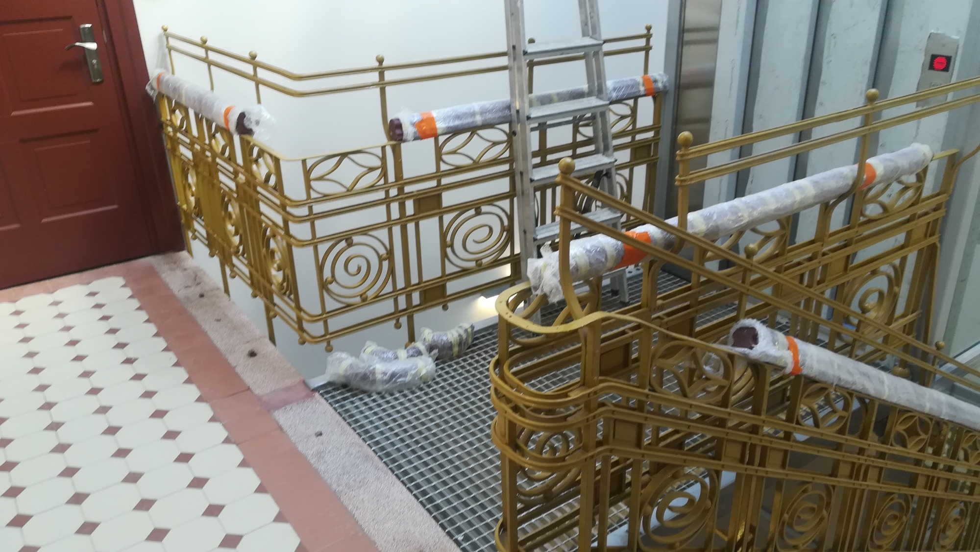 Złote, metalowe balustrady schodowe z dekoracyjnymi elementami spiralnymi, częściowo owinięte folią ochronną, ustawione na metalowej kracie, z aluminiową drabiną opartą o balustradę, w tle brązowe...