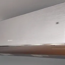 Nowoczesny srebrny klimatyzator marki Gree zamontowany na szarej ścianie, widoczne detale obudowy i logo producenta.