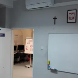 Biały klimatyzator ścienny Mitsubishi w klasie szkolnej, widoczny nad biurkiem nauczycielskim z dekoracjami, krzyż i godło Polski na ścianie.