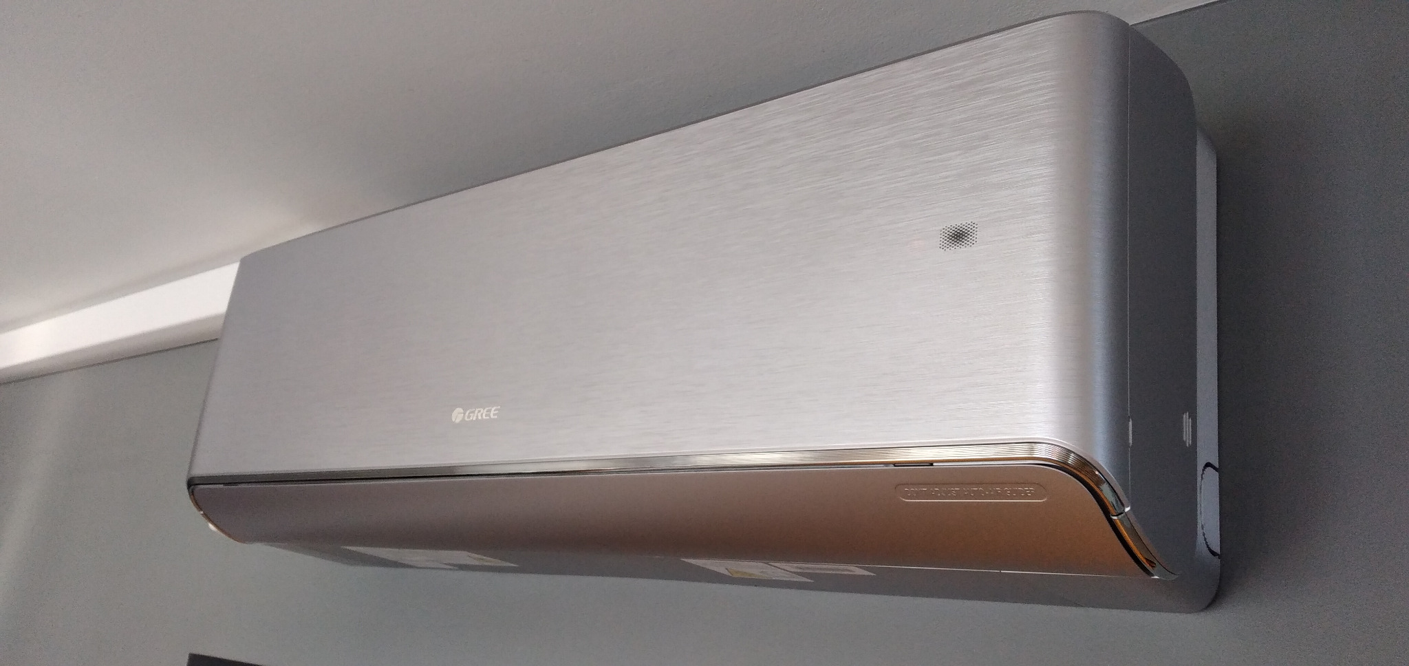 Nowoczesny srebrny klimatyzator marki Gree zamontowany na szarej ścianie, widoczne detale obudowy i logo producenta.