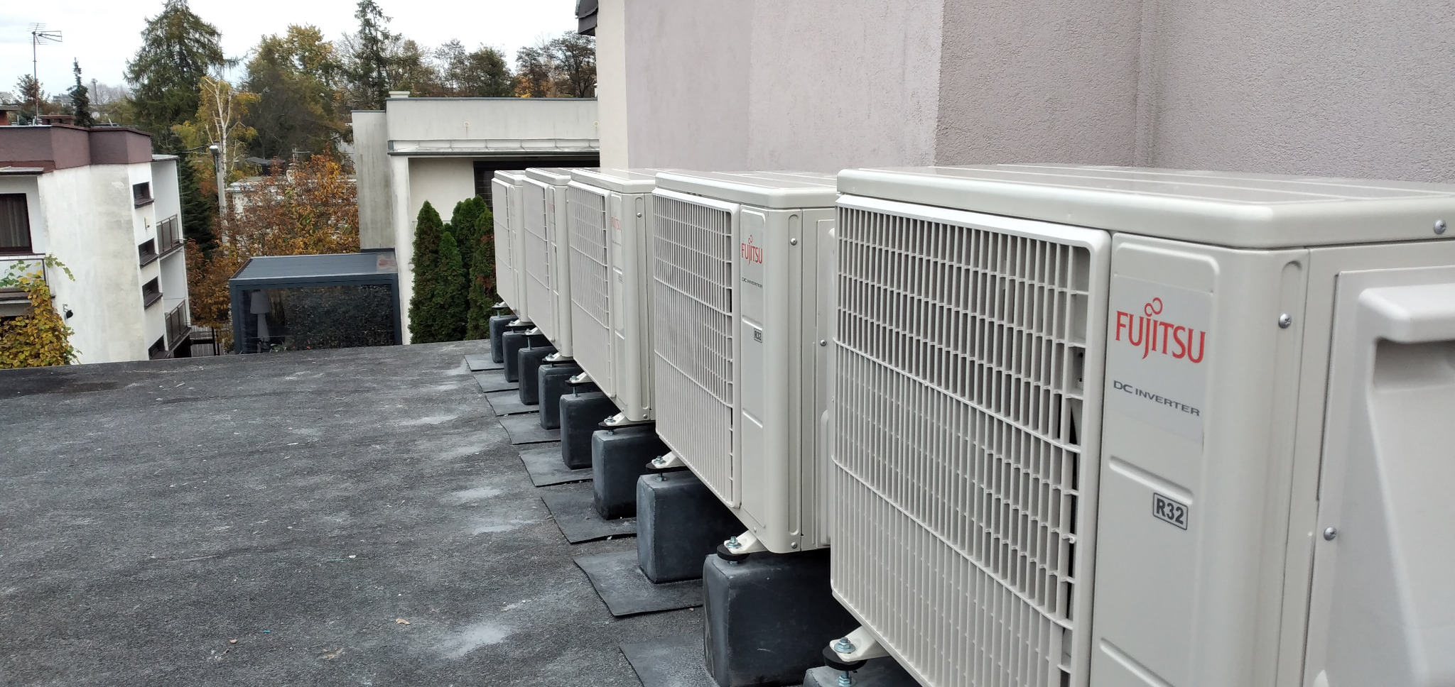 Rząd klimatyzatorów Fujitsu DC Inverter R32 na dachu budynku, zamontowanych na betonowych podstawach, z widokiem na drzewa i inne budynki w tle.