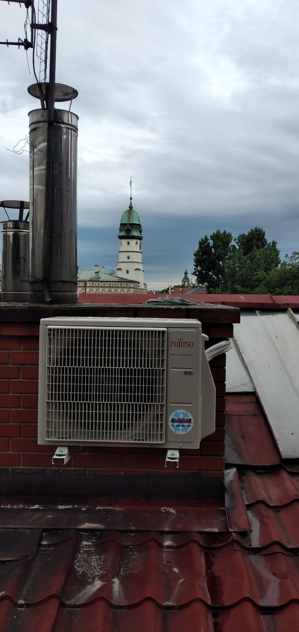 Zewnętrzna jednostka klimatyzacji Fujitsu zamontowana na ceglanym kominie na dachu z czerwonej dachówki, w tle wieża ratuszowa i pochmurne niebo.