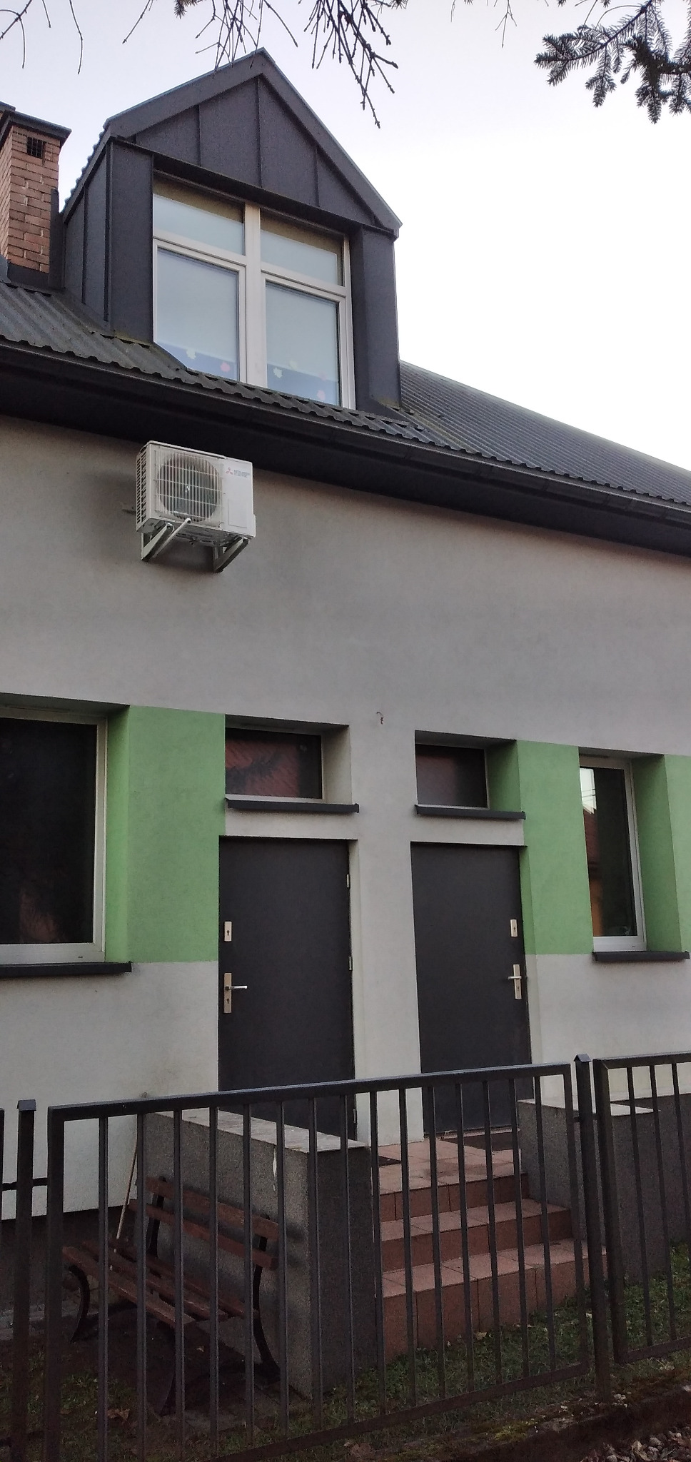 Zewnętrzna jednostka klimatyzacji Mitsubishi Electric zamontowana na ścianie budynku z dwoma ciemnymi drzwiami i oknami, widok zza metalowego ogrodzenia.