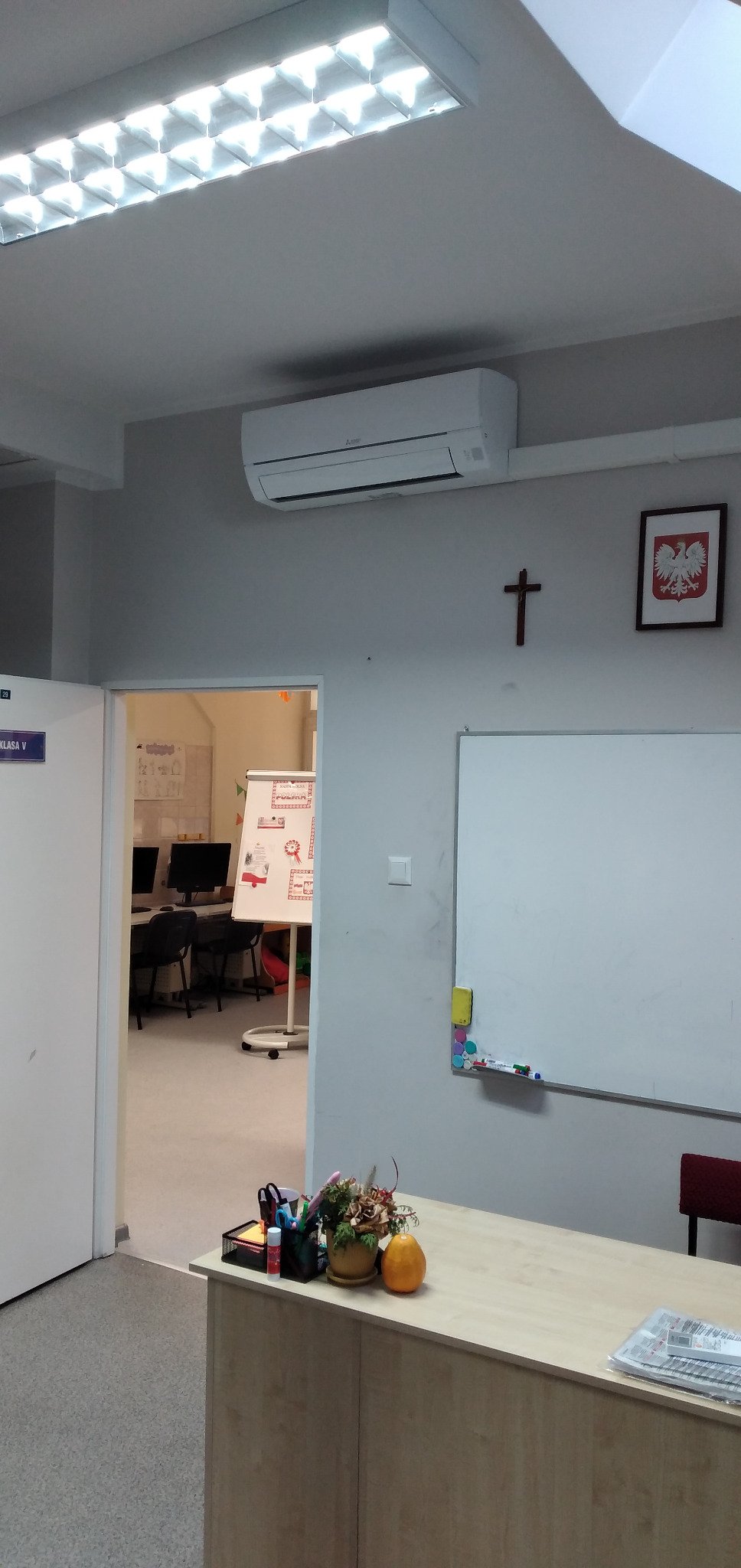 Biały klimatyzator ścienny Mitsubishi w klasie szkolnej, widoczny nad biurkiem nauczycielskim z dekoracjami, krzyż i godło Polski na ścianie.