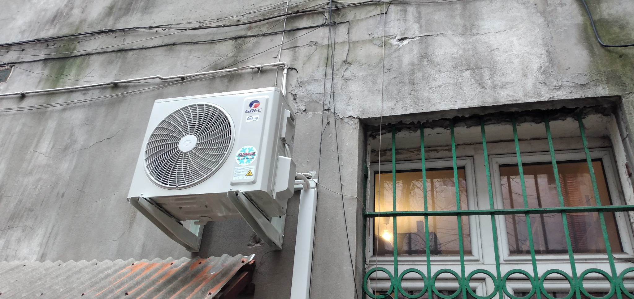 Zewnętrzna jednostka klimatyzacji Gree zamontowana na elewacji budynku z widocznymi kablami i oknem z zielonymi kratami.