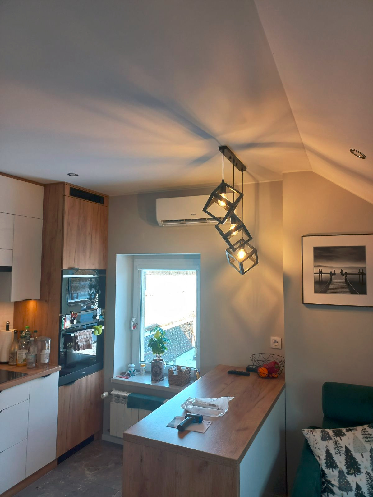 Wnętrze kuchni z zamontowanym klimatyzatorem ściennym marki Gree, widocznym nad oknem, oraz nowoczesną lampą wiszącą z trzema kwadratowymi kloszami. Na blacie fragment instalacji.
