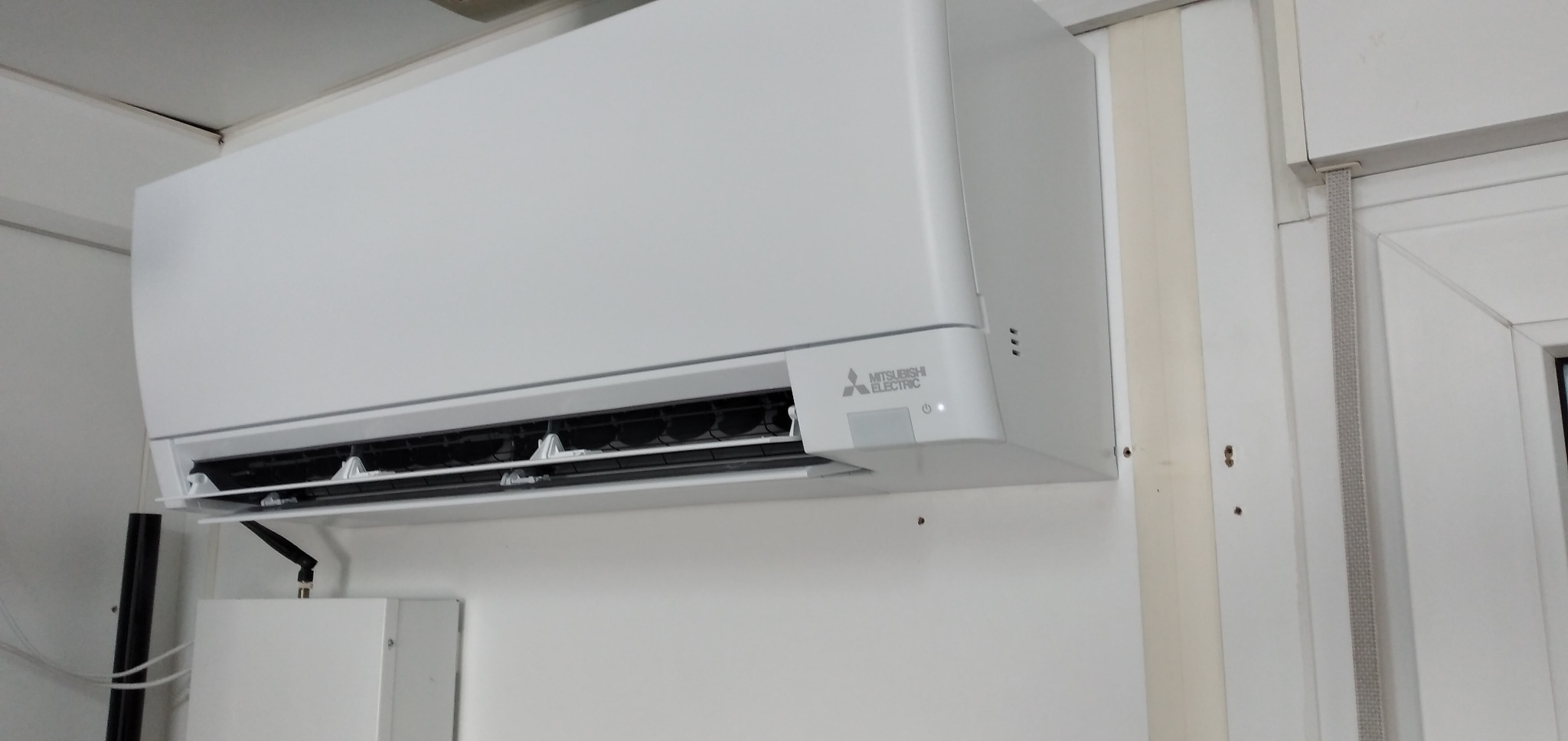 Biała jednostka wewnętrzna klimatyzacji Mitsubishi Electric zamontowana na białej ścianie, widoczne otwory wentylacyjne i panel sterowania z logo.