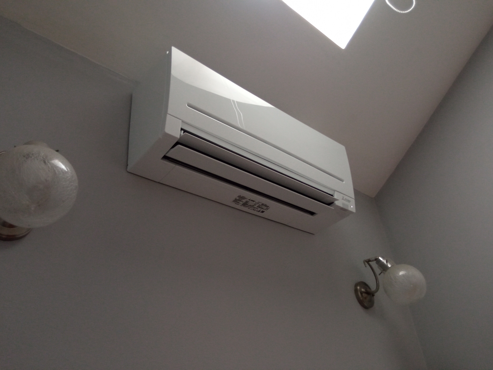 Biały klimatyzator Mitsubishi zamontowany na szarej ścianie, widoczne dwa kinkiety z mlecznymi kloszami i fragment sufitu z oświetleniem.