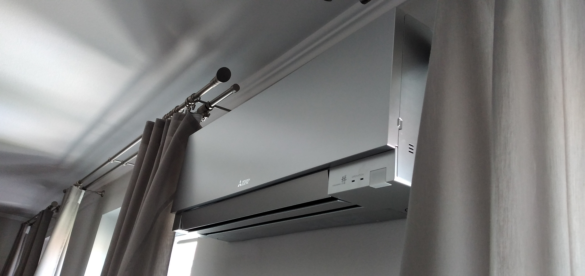 Szary klimatyzator Mitsubishi Electric zamontowany w domu, widoczny z dołu, obok zasłon na podwójnym karniszu.