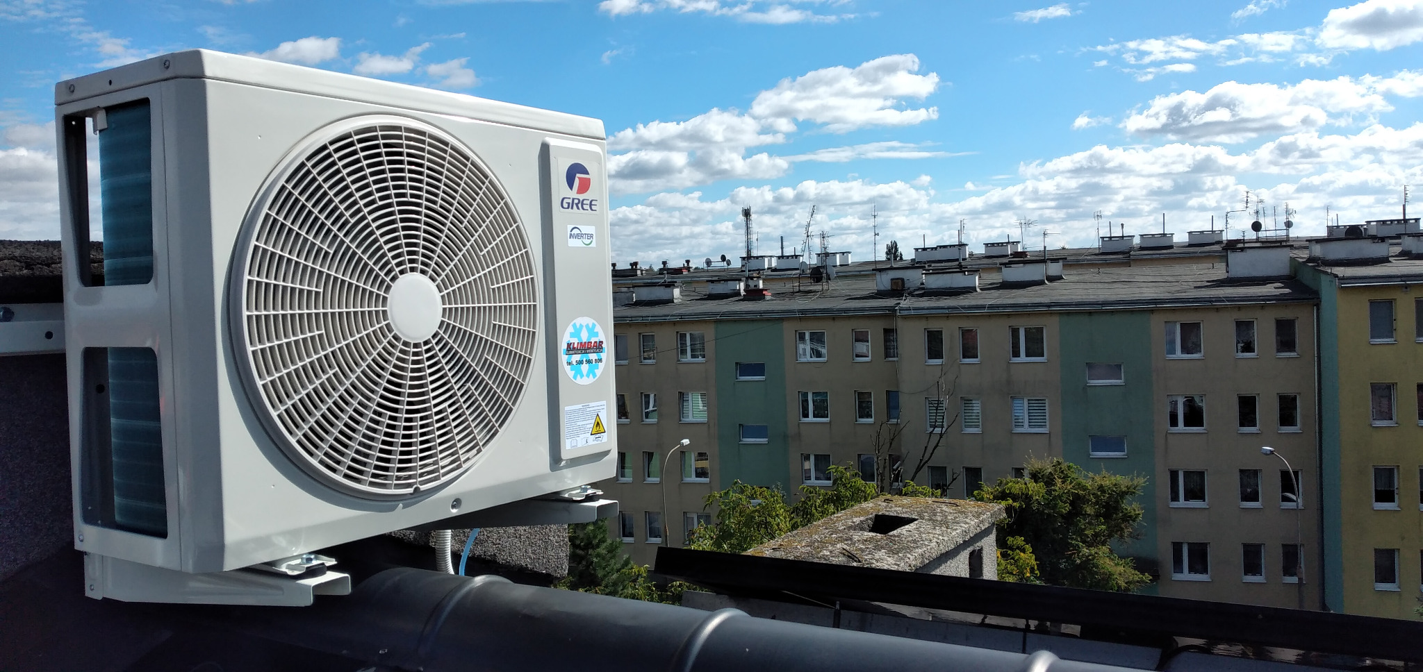 Zewnętrzna jednostka klimatyzatora marki Gree zamontowana na dachu budynku wielorodzinnego, widoczne anteny i kominy na dachu, błękitne niebo z chmurami w tle.