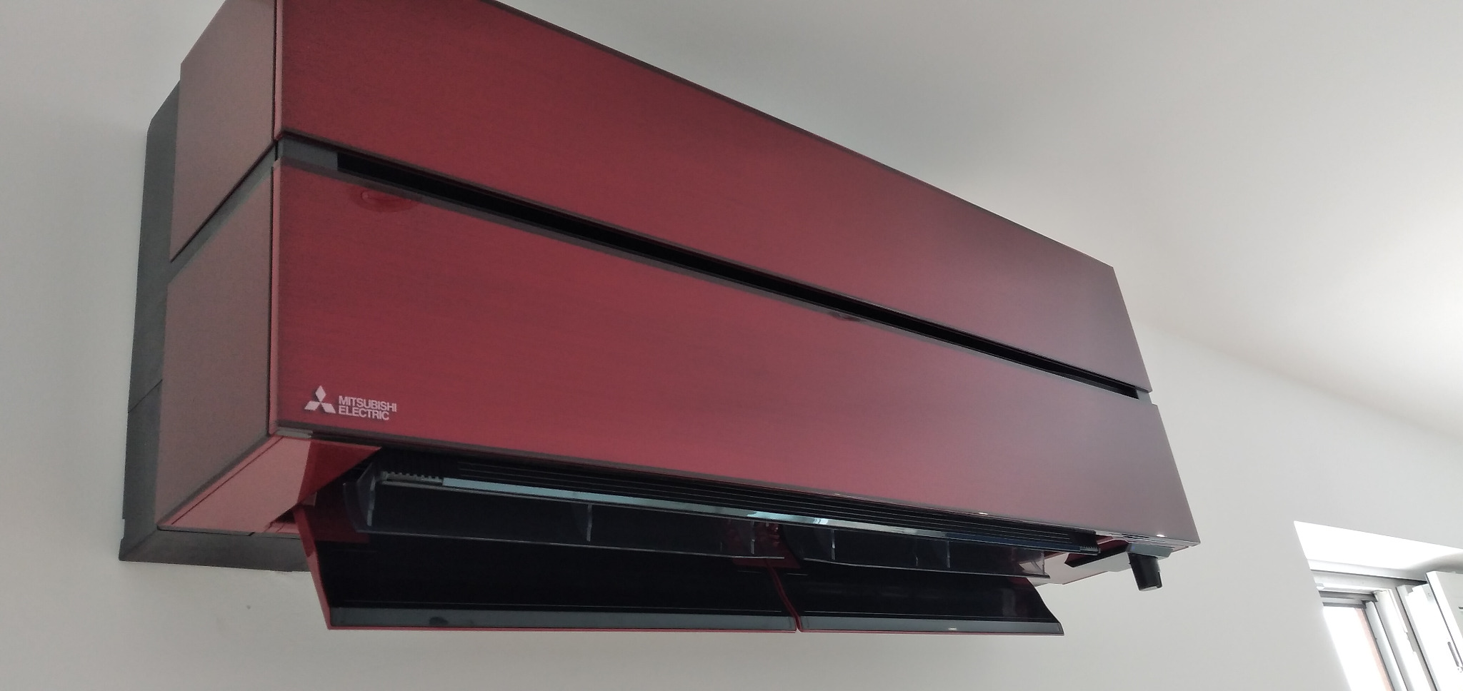 Czerwony klimatyzator Mitsubishi Electric zamontowany na białej ścianie, widoczne otwarte żaluzje i logo producenta.