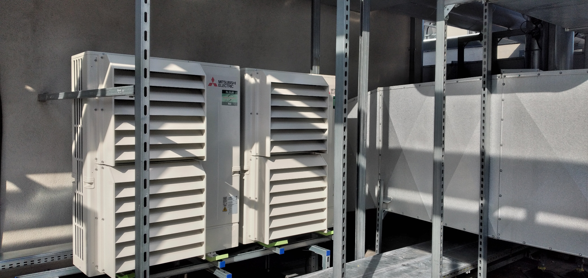 Dwa moduły zewnętrzne klimatyzacji Mitsubishi Electric zamontowane na konstrukcji wsporczej na dachu budynku, widoczne elementy wentylacyjne i kanały powietrzne.