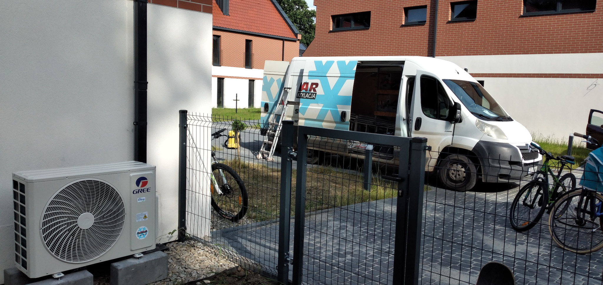 Zewnętrzna jednostka klimatyzacji GREE zamontowana przy ścianie domu, obok siatki ogrodzeniowej, z dostawczym busem firmy klimatyzacyjnej w tle i rowerem opartym o ogrodzenie.