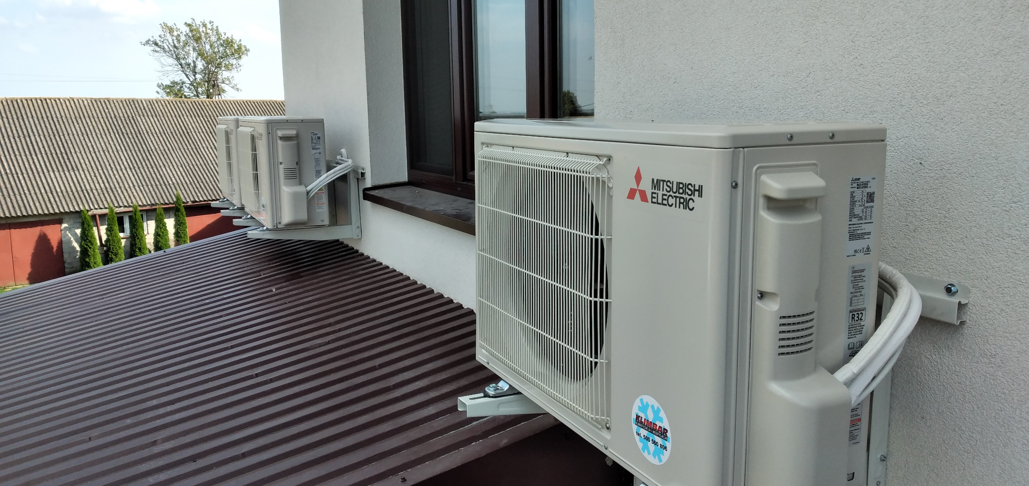 Dwa klimatyzatory Mitsubishi Electric zamontowane na zewnątrz budynku, widoczne przewody i uchwyty montażowe, na jednym z urządzeń naklejka firmy serwisującej.