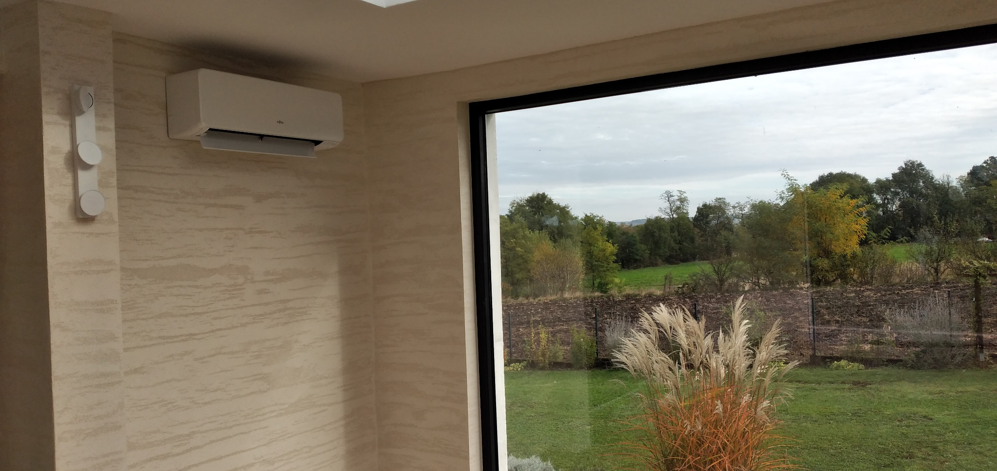 Nowoczesny klimatyzator marki Fujitsu zamontowany na ścianie z fakturą, widok przez duże okno na zewnątrz z ozdobnymi trawami.