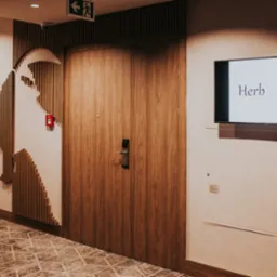 Dwuskrzydłowe drzwi wewnętrzne z drewnianą okleiną w korytarzu hotelowym, obok dekoracyjna instalacja ścienna z drewnianych listew i telewizor z logo 'Herb'.