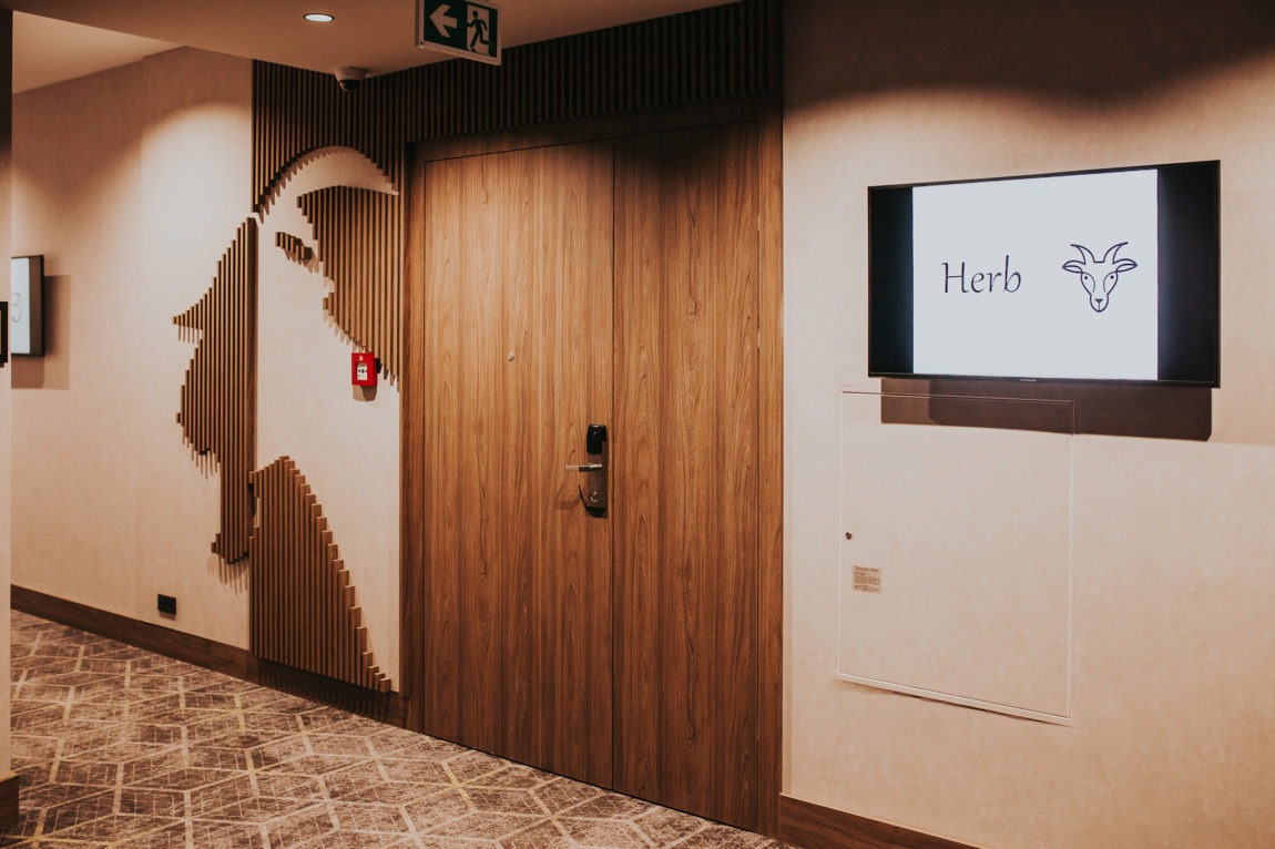 Dwuskrzydłowe drzwi wewnętrzne z drewnianą okleiną w korytarzu hotelowym, obok dekoracyjna instalacja ścienna z drewnianych listew i telewizor z logo 'Herb'.