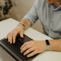 Mikołaj Londo Coco Bystre Rozwiązania - Mężczyzna w jasnej koszuli pisze na laptopie. Na nadgarstku zegarek sportowy, na palcu obrączka. Widoczne dłonie na klawiaturze i fragment twarzy.