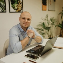 Mikołaj Londo Coco Bystre Rozwiązania - Mężczyzna w okularach siedzi przy biurku z laptopem, telefonem i długopisem. W tle rośliny i obrazy w ramkach. Ubrany w błękitną koszulę, z bransoletką na ręce.