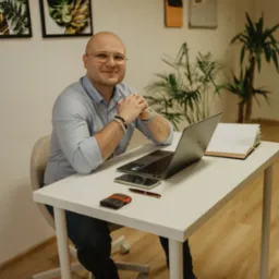 Mężczyzna w okularach siedzi za biurkiem z laptopem, telefonem i notatnikiem. W tle widoczne rośliny i dekoracje ścienne, tworzące przyjazną atmosferę.