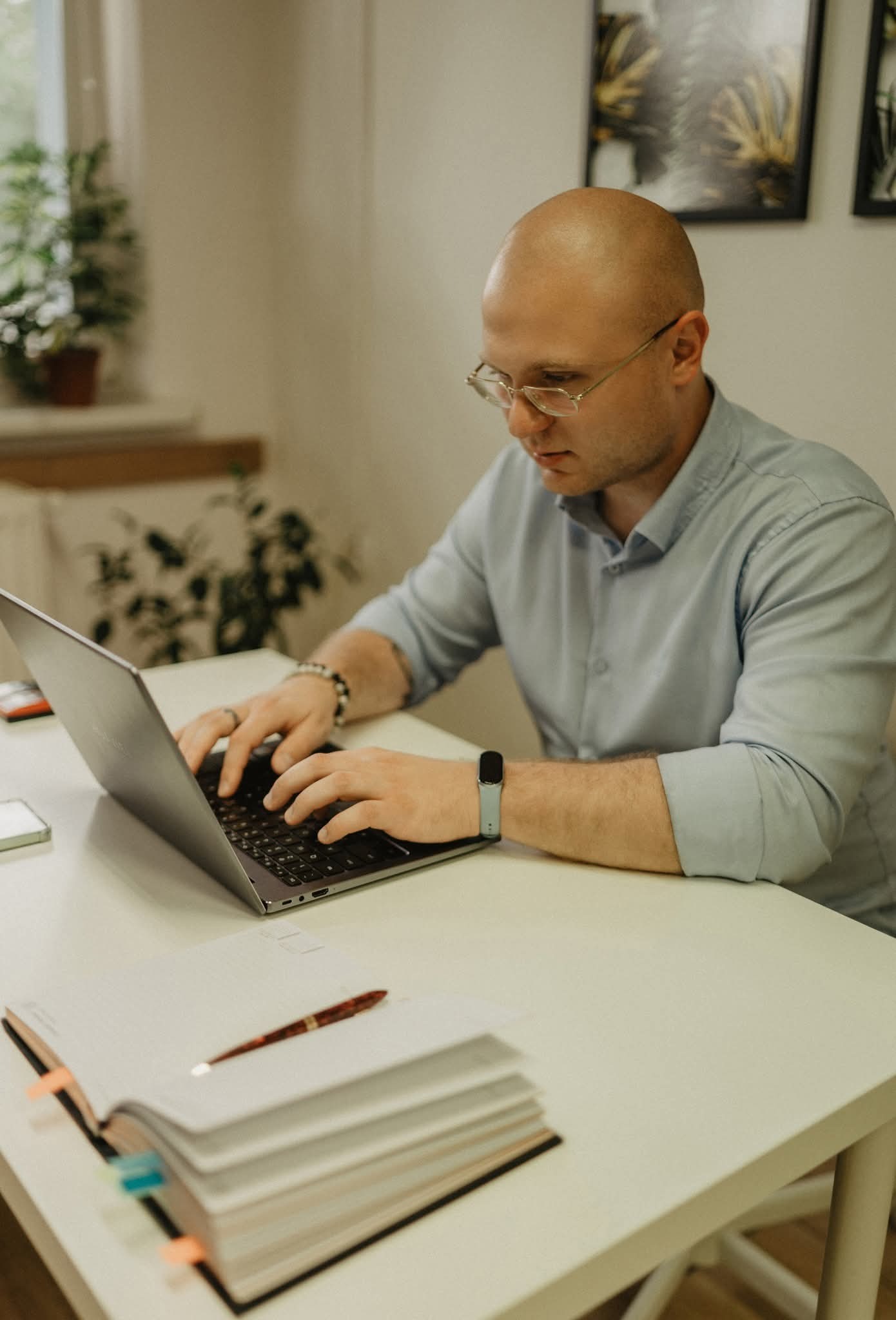 Mężczyzna w okularach skupiony na pracy przy laptopie. Na stole otwarty notatnik z zakładkami, obok leży pióro. W tle roślina i grafiki na ścianie.