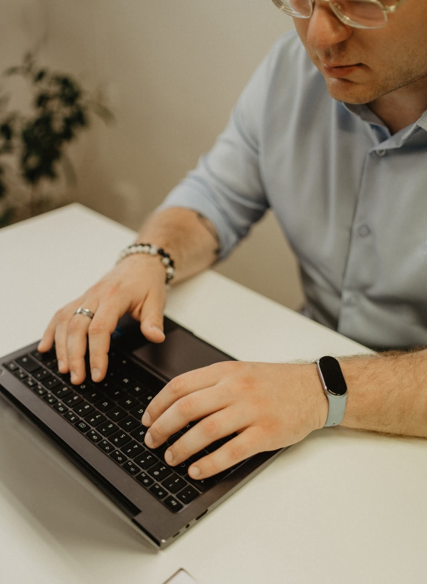 Mężczyzna w jasnej koszuli pisze na laptopie. Na nadgarstku zegarek sportowy, na palcu obrączka. Widoczne dłonie na klawiaturze i fragment twarzy.