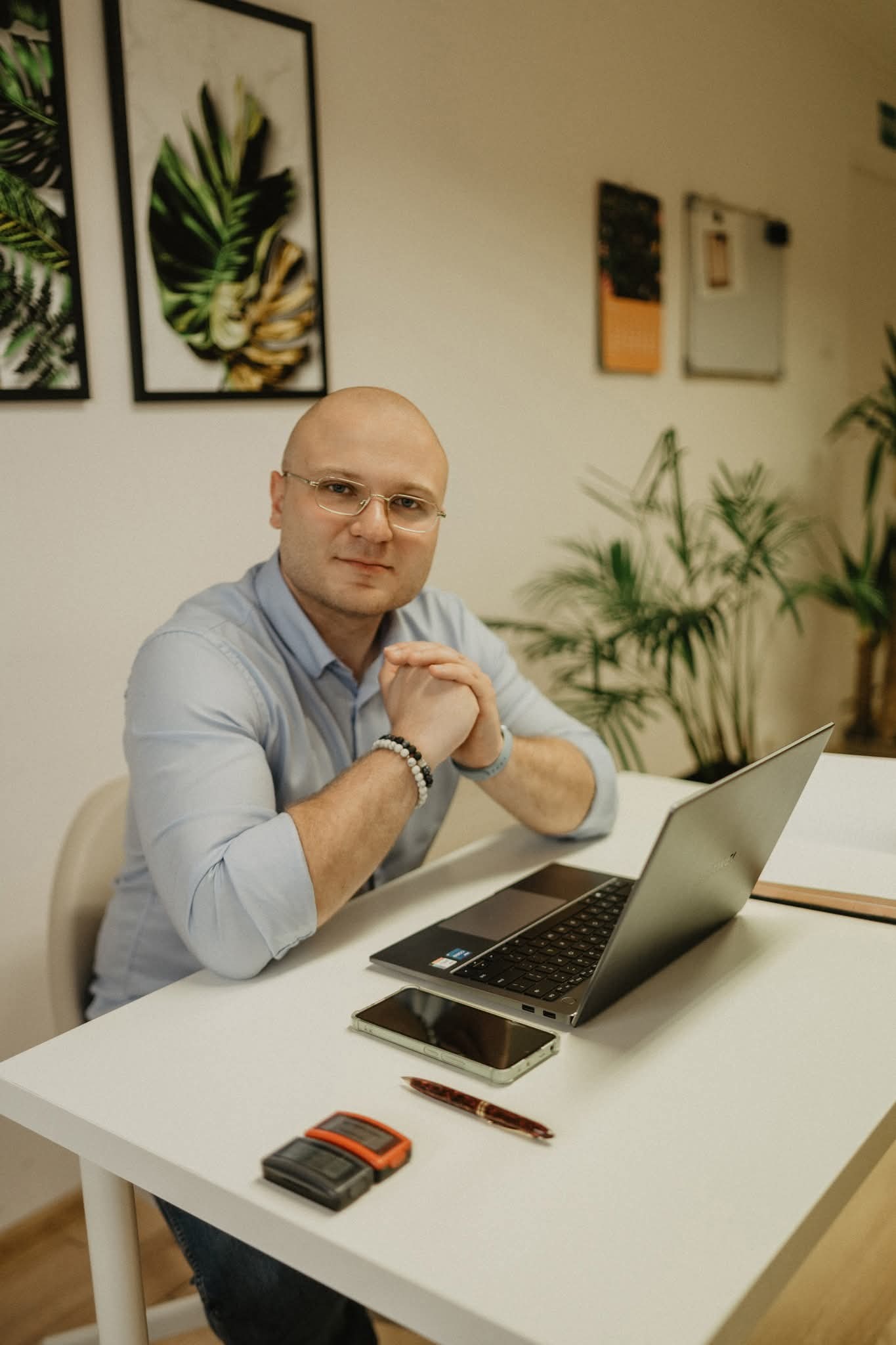 Mężczyzna w okularach siedzi przy biurku z laptopem, telefonem i długopisem. W tle rośliny i obrazy w ramkach. Ubrany w błękitną koszulę, z bransoletką na ręce.