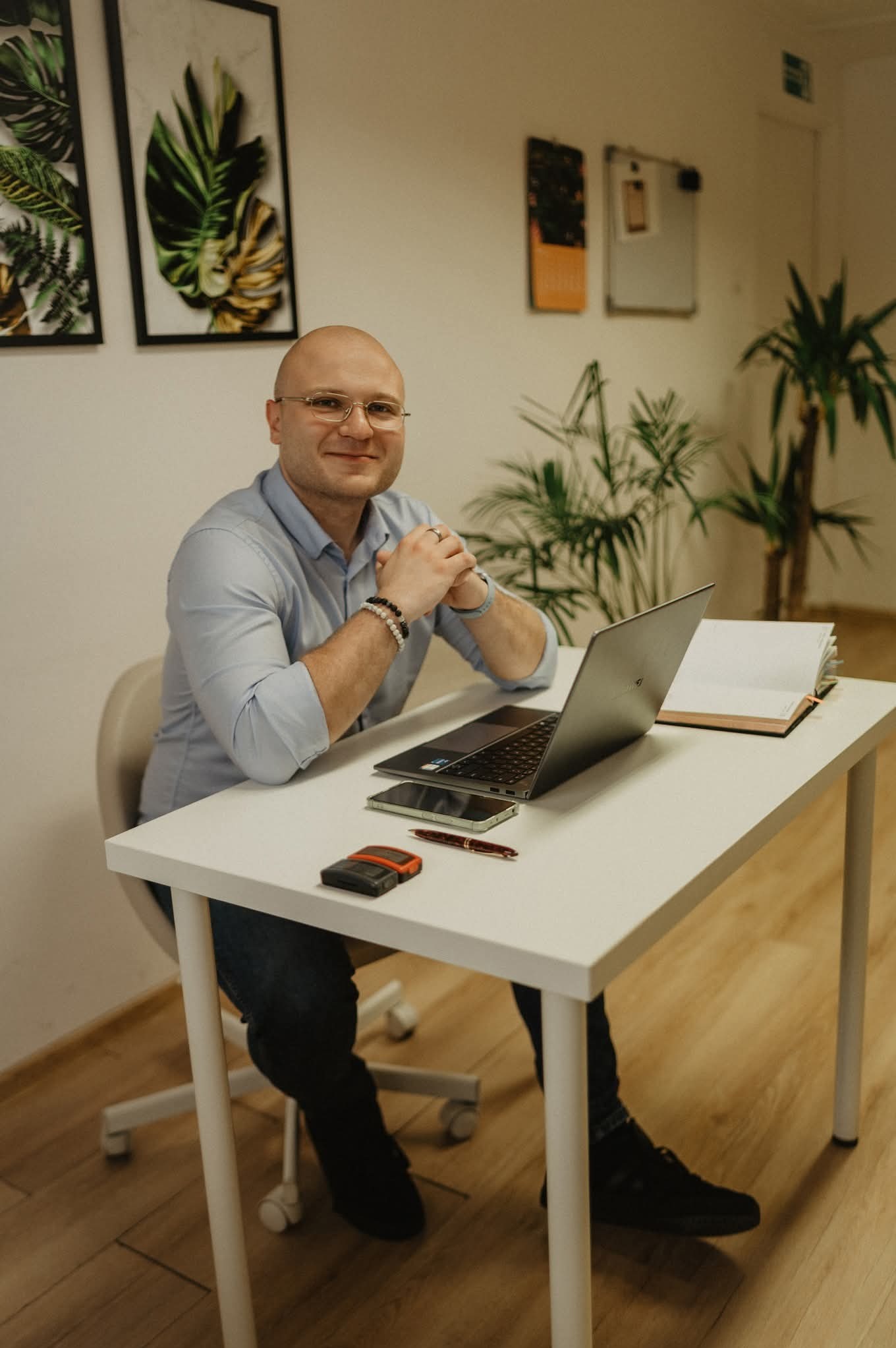 Mężczyzna w okularach siedzi za biurkiem z laptopem, telefonem i notatnikiem. W tle widoczne rośliny i dekoracje ścienne, tworzące przyjazną atmosferę.