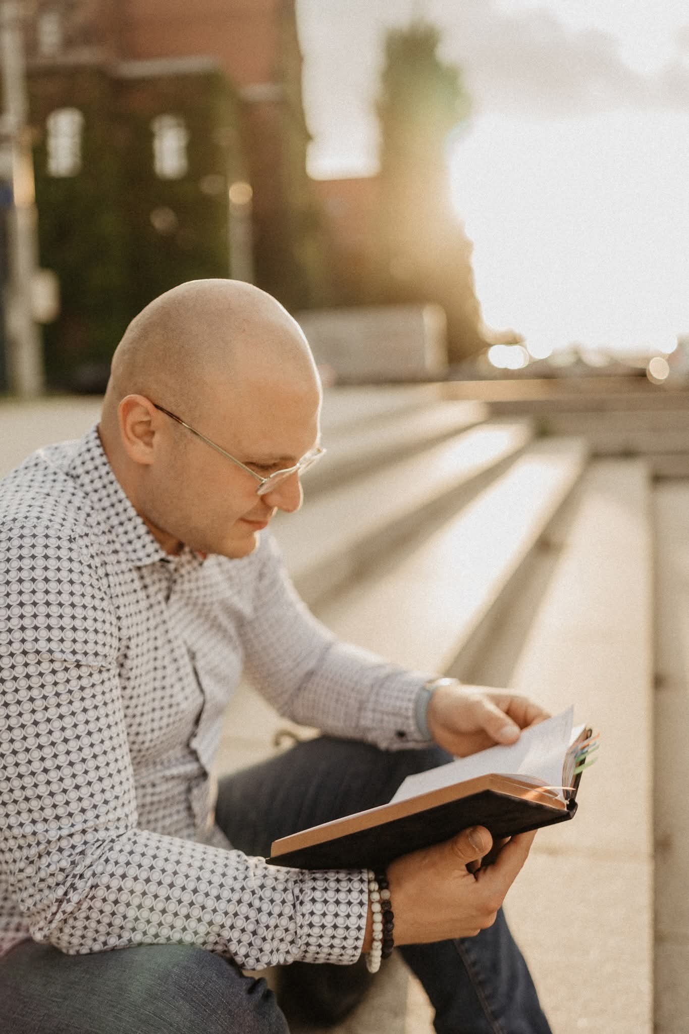 Mężczyzna w okularach, siedząc na schodach w popołudniowym słońcu, przegląda książkę z zakładkami. Ubrany w koszulę w geometryczny wzór i jeansy.