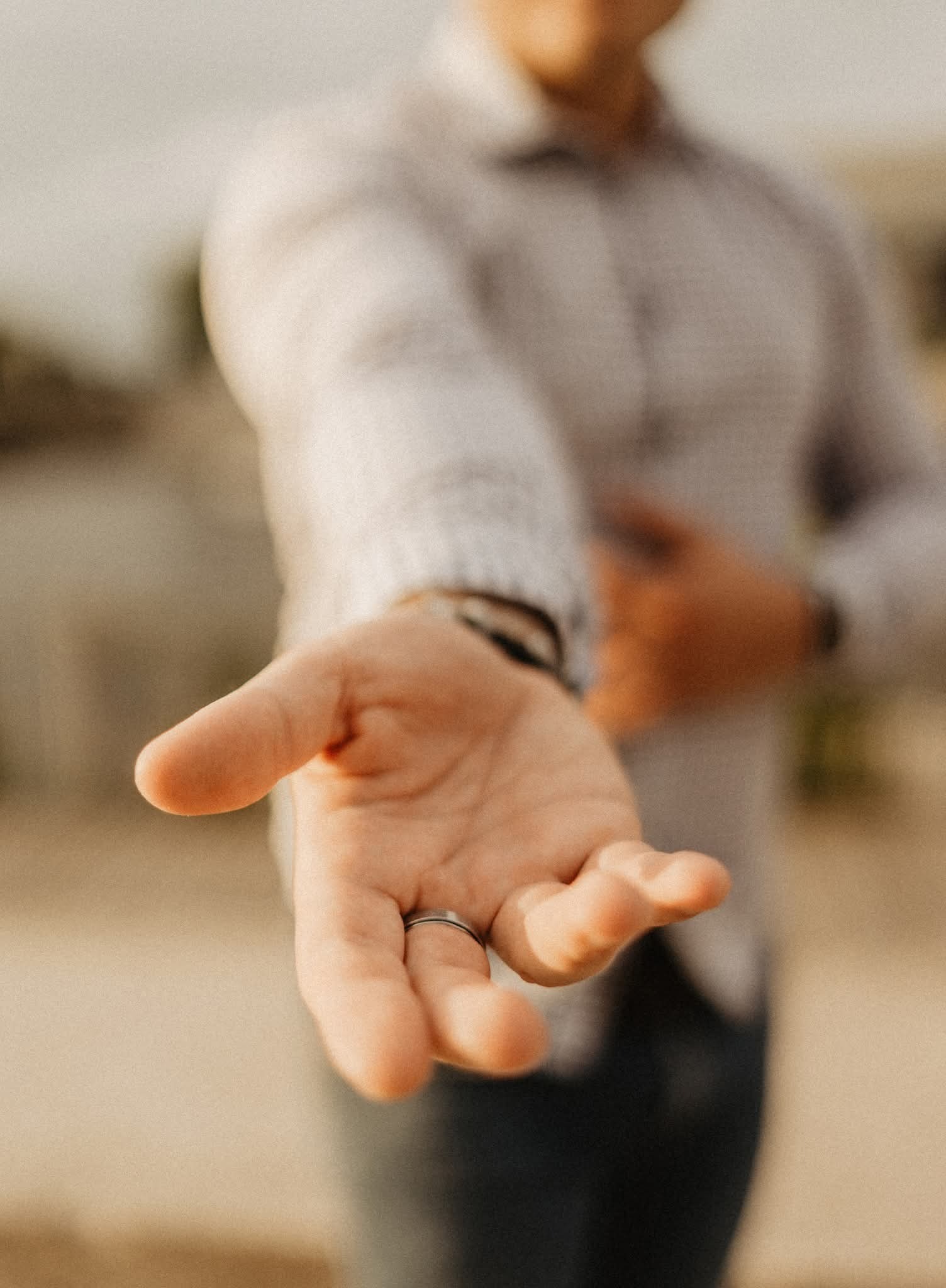 Otwarta dłoń z pierścionkiem, wyciągnięta w geście zaproszenia lub pomocy, na rozmytym tle osoby w koszuli, z delikatnym, naturalnym oświetleniem.