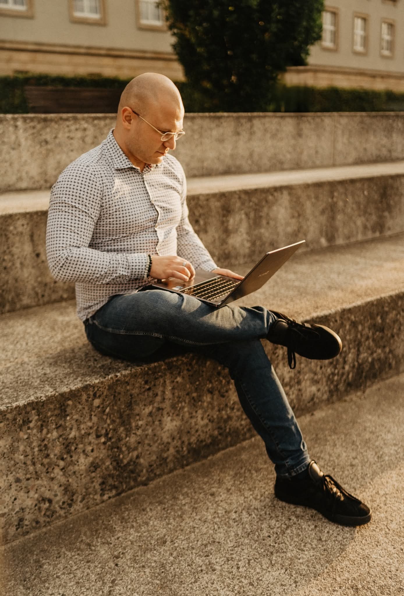 Mężczyzna w okularach, siedząc na betonowych schodach w promieniach słońca, pracuje na laptopie. Ubrany w wzorzystą koszulę i jeansy. Tło stanowi budynek i zieleń.