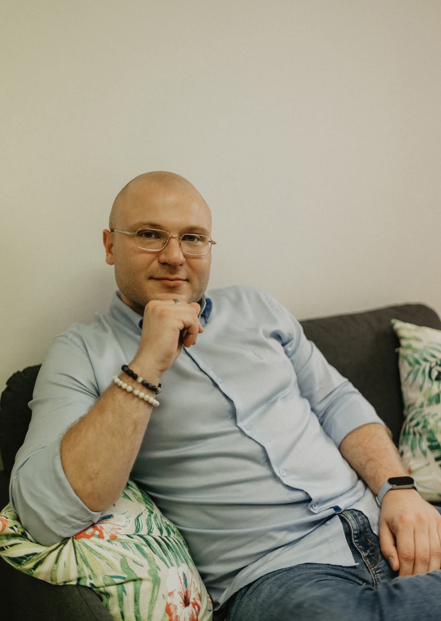 Portret mężczyzny w okularach, siedzącego na szarej kanapie z poduszkami w tropikalne wzory. Ma na sobie błękitną koszulę i dżinsy. Na rękach widoczne bransoletki z kamieni.