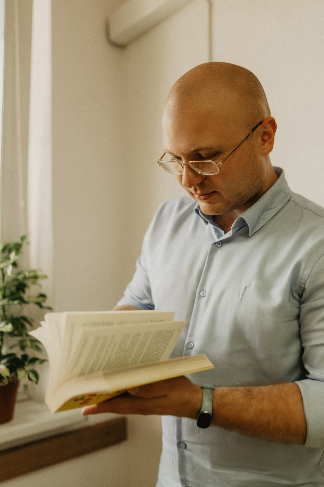 Mężczyzna w okularach czyta książkę przy oknie, obok doniczka z rośliną. Ubrany w błękitną koszulę. Delikatne światło pada na twarz.